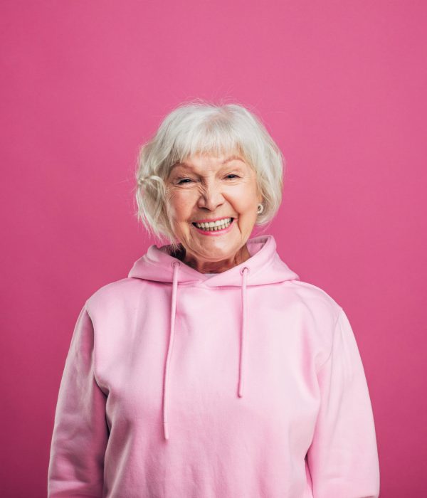 Happy cheerful positive old woman smiling wide and look straight on camera. Stand straight. Modern stylish senior model with grey hair. Isolated over pink background